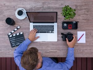 A man using a laptop holding a camera with some recording equipment around him.